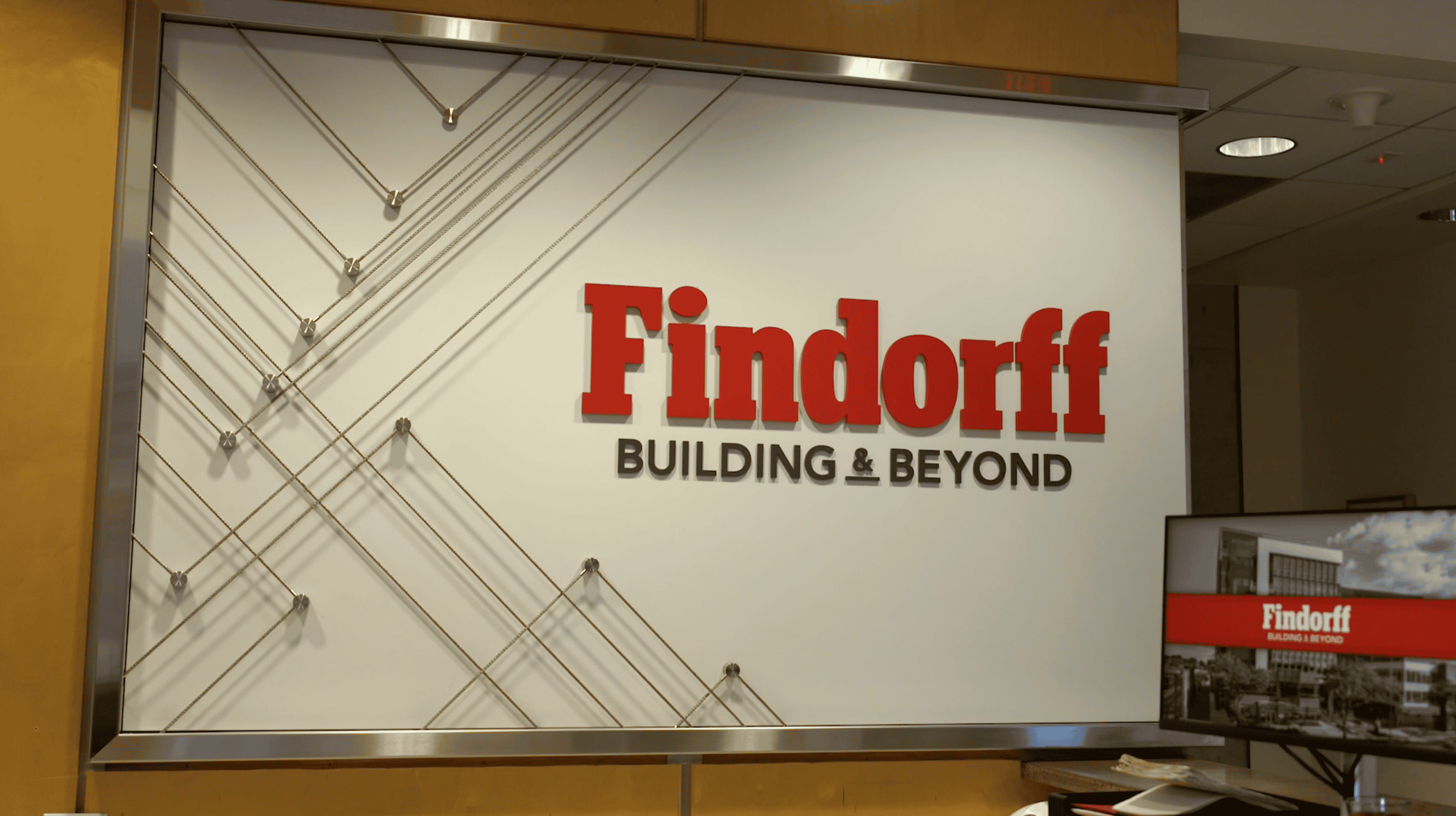 A branded wall installation displaying the Findorff logo with the tagline "Building & Beyond" in bold red and black letters. The logo is set against a white background, framed by metal with a pattern of intersecting diagonal cables. The installation is located in a modern office lobby with warm wood paneling, part of a facility branding project.